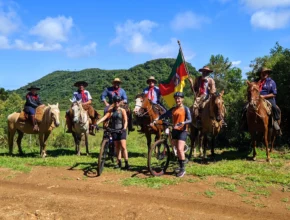 Grupo de tropeiros renascentes a cavalo e cicloturistas contemporâneos no Costão do Cambará, Campos de Cima da Serra (RS), com bandeira histórica e paisagem montanhosa. Foto: Ivan Mendes © Lobi Ciclotur