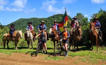 Grupo de tropeiros renascentes a cavalo e cicloturistas contemporâneos no Costão do Cambará, Campos de Cima da Serra (RS), com bandeira histórica e paisagem montanhosa. Foto: Ivan Mendes © Lobi Ciclotur