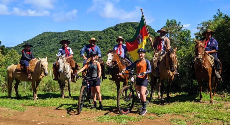 Grupo de tropeiros renascentes a cavalo e cicloturistas contemporâneos no Costão do Cambará, Campos de Cima da Serra (RS), com bandeira histórica e paisagem montanhosa. Foto: Ivan Mendes © Lobi Ciclotur